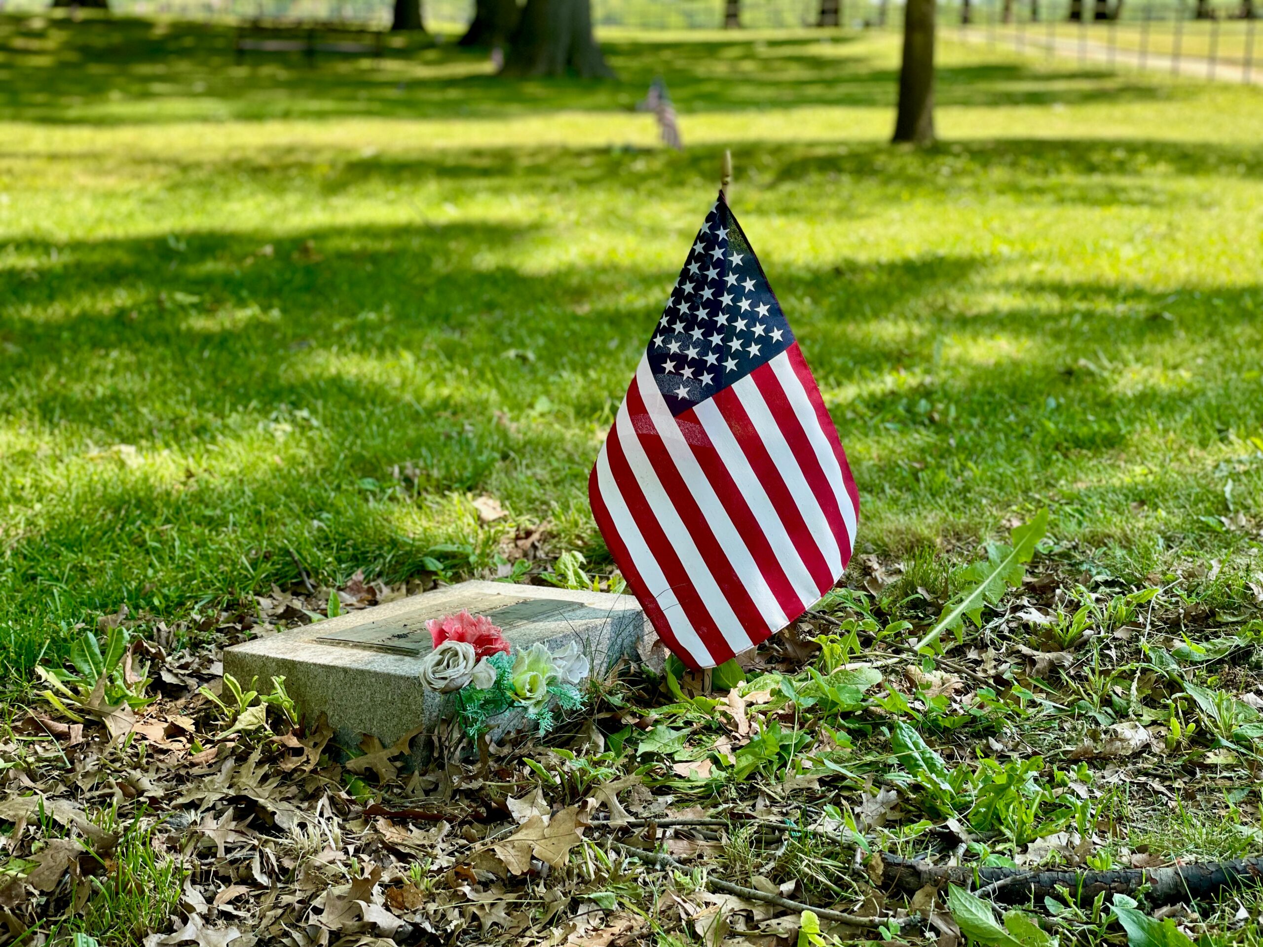 A solemn scene with an American flag beside a tombstone in a grassy cemetery.